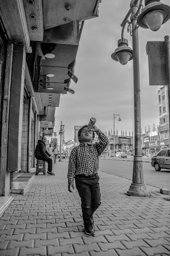 Grayscale Photo of Boy in Dress Shirt Walking on Side Walk While Drinking from a Glass Bottle
