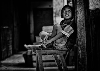 grayscale photo of boy sitting on wooden bench beside wall while looking up