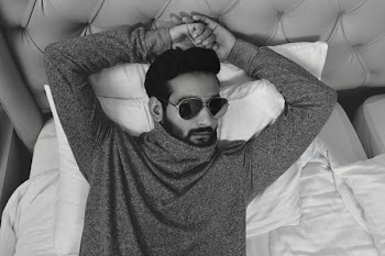 Grayscale Photo of Man in Long-sleeved Wearing Sunglasses Lying on Bed With Both Hand on Head