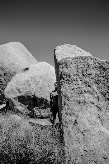Grayscale Photo of Man Standing Besides Large Rock
