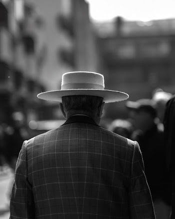 grayscale photo of man wearing hat standing beside building