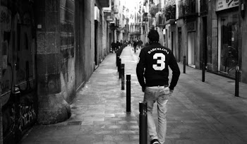 Grayscale Photo of Man Wearing Long-sleeved Shirt and Pants Walking on Concrete Pathway Near Building