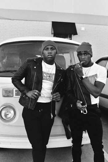 grayscale photo of two men wearing jacket while standing infront of vehicle
