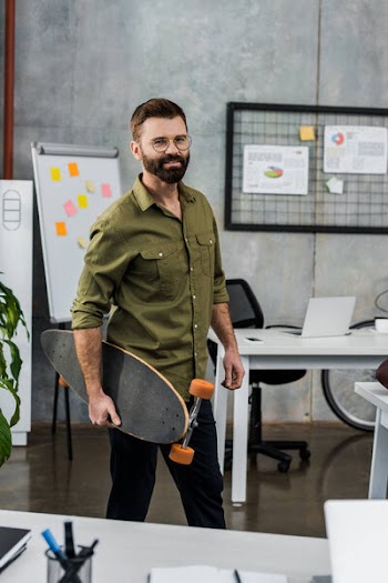 Handsome bearded businessman in eyeglasses holding skateboard and smiling at camera in office