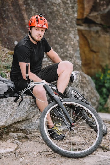 Handsome young trial biker relaxing on rocks with bicycle outdoors and looking at camera