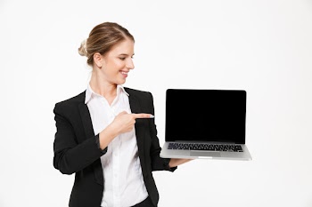 Happy blonde business woman showing blank laptop computer screen while looking and pointing at him over white wall Free Photo
