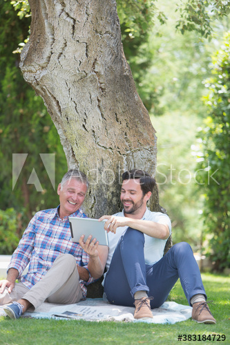 Happy father and adult son using digital tablet below tree in park