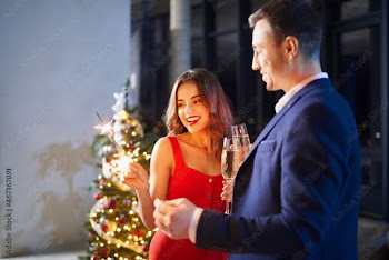 Happy man and woman holding sparkles and glass of champagne while relaxing together during evening time. Young family celebrating winter holidays ay home.