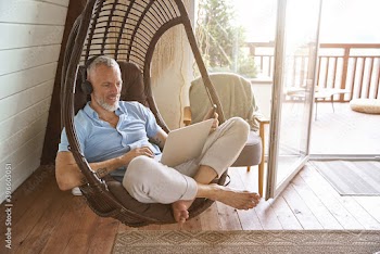 Happy middle aged caucasian businessman wearing headphones using laptop while sitting on swing chair at his modern apartment and working from home