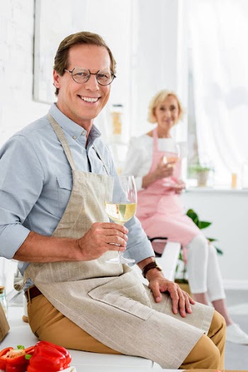 Happy senior couple holding glasses of wine and smiling at camera while cooking together