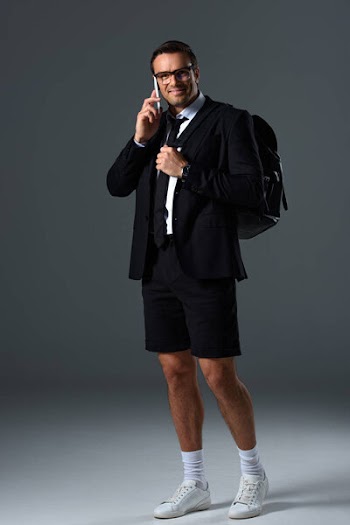 Happy stylish man in shorts with backpack talking on smartphone on grey background