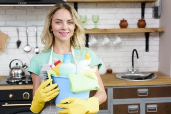 Happy young housekeeper standing in kitchen holding bucket of cleaning products Free Photo