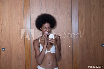 Happy young woman with afro in bra using smart phone at closet