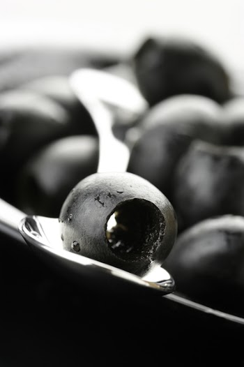 Heap of black olives in plate and olive in spoon close-up.