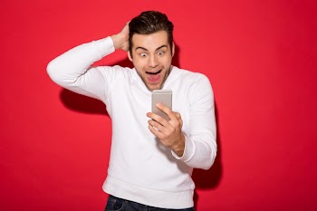 Image of surprised happy man in sweater looking at smartphone over red wall Free Photo