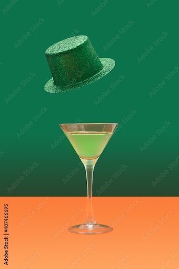 Irish festival minimal concept. Martini glass with green cocktail on terracotta table. Green glitter hats levitate above on a dark green background.