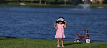 Kid in Pink Dress With White and Black Sun Bucket Hat Beside the Red Pedal Trike in Front of Body of Water
