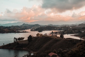 Landscape Photography of Houses on Green Field Viewing Mountain and Body of Water Under White and Orange Sky