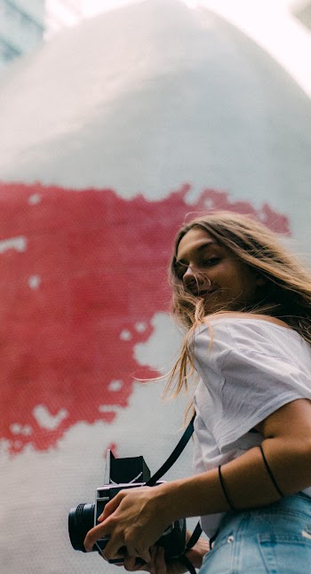 Low Angle Photo of Smiling Woman in White T-shirt Standing While Carrying Black Dslr Camera