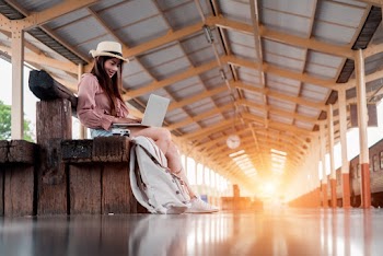 Low Angle Photo of Smiling Woman Using A Laptop Sitting on Wooden Bench at Train Station Platform
