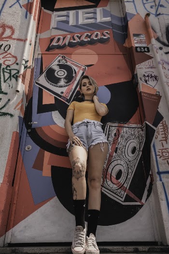 Low Angle Photo of Tattooed Woman in Orange T-shirt and Blue Denim Shorts Leaning on Graffiti Door