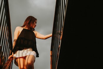 Low Angle Photo of Woman in Black Top and White Skirt Looking Away While Standing at the top of a Staircase Holding Metal Railing