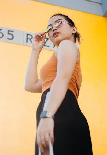 Low Angle Photo of Woman in Sunglasses,Orange Top, and Black Bottoms Standing Beside Yellow Wall