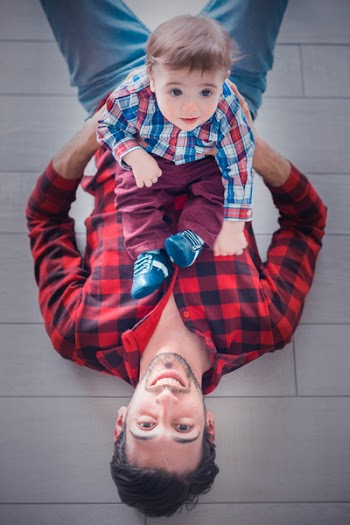 Lying Man Wearing Red and Black Gingham Long-sleeved Shirt Holding Toddler Sitting on His Stomach
