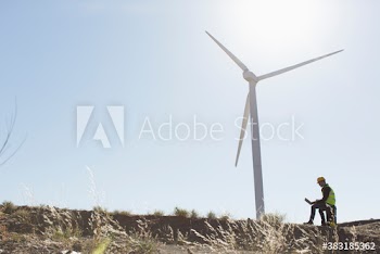 Male engineer with laptop working below sunny wind turbine