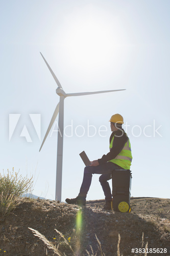 Male engineer with laptop working below sunny wind turbine