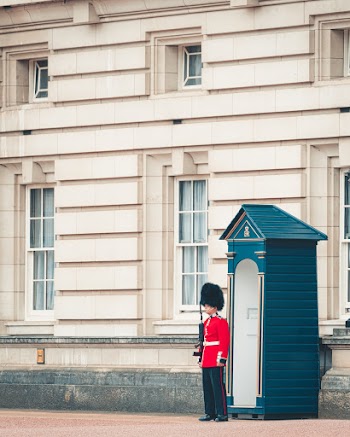 Male Guard Wearing Red and White Costume Holding Rifle While Standing Ear White and Blue Shed