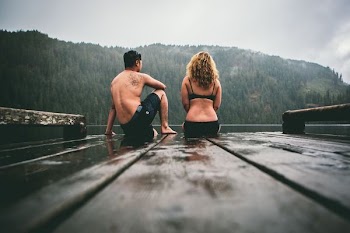 man and woman sitting on brown wooden boat dock facing body of water under gray skies