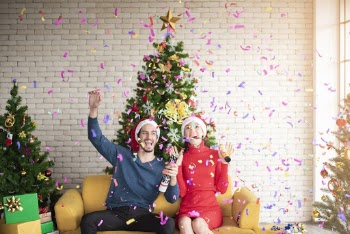 Man and Woman Wearing Santa Hats Sitting on Sofa Popping A Confetti Surrounded By Christmas trees
