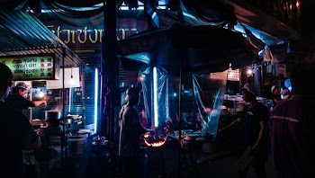 man grilling meat while man in black shirt standing in front of food stall