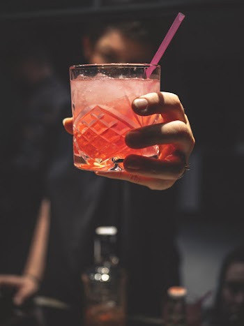 Man Holding Forward A Glass Of Iced Pinkish Colored Cocktail With A Straw