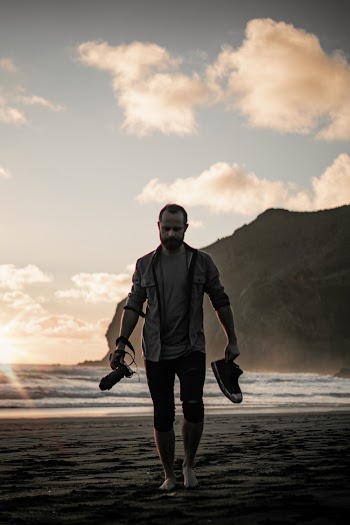 Man Holding Shoes And Camera Walking At The Beach