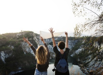 Man Holding White Pennant Beside Girl Raising Two Hands in Front of Lake Surrounded by Mountains