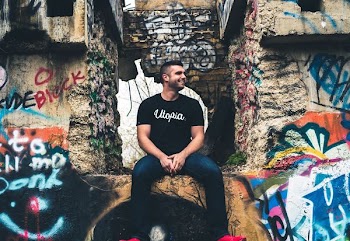 Man in a t-shirt reading "Utopia" smiles to the side while sitting among concrete graffitied walls