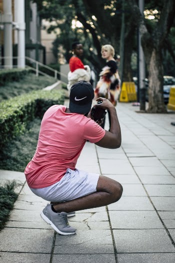 Man In Black Cap, Pink T-shirt, White Shorts, And Grey Shoes Holding Camera