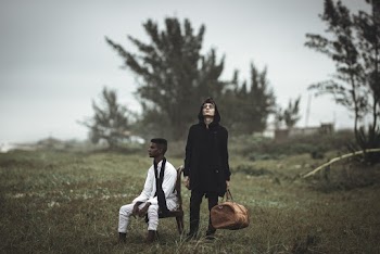 Man in Black Coat Standing Beside Man Sitting on Chair Wearing White Clothes 