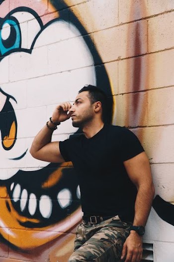 man in black crew-neck shirt and green and beige camouflaged pants leaning on wall with lola bunny graffiti