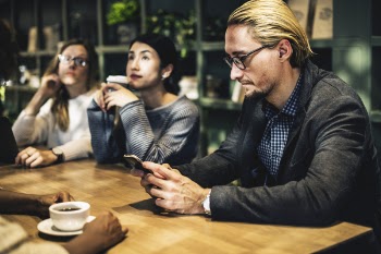 Man in Black Suit Jacket Holding Smartphone Sitting in Front of Table