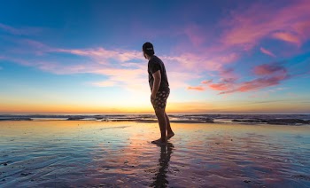 Man in Black T-shirt Wearing Cap Near Body of Water during Sunset