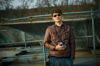 Man in Brown Leather Jacket Wearing Black Framed Sunglasses Holding Black Dslr Camera Standing in Front of Black Steel Bar during Daytime