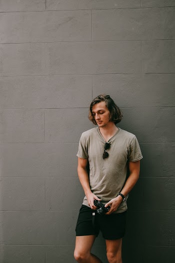 Man in Gray Tee Shirt Holding Camera While Leaning on Wall