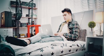 Man in Green, Blue, and Black Plaid Sports Shirt Sitting on Bed Using Silver Macbook