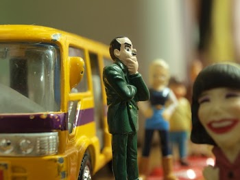 Man in Green Coat Figure Standing in Front of Yellow Toy Bus