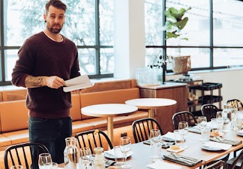 Man in Maroon Sweater Standing Beside Table Holding Book