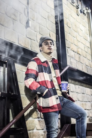 Man in Red, Black, and Grey Plaid Button-up Jacket With Blue Denim Jeans Outfit Holding Disposable Cup Stands Beside Brown Concrete Wall