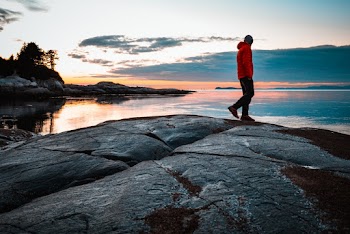 Man in Red Jacket Standing on Stone Looking at Body of Water